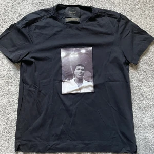 Svart t-shirt Limitato  - Svart t-shirt från Limitato med ikoniskt tryck av Muhammad Ali på bröstet. Klassisk passform med rund hals och korta ärmar. Tillverkad i mjuk bomull som känns skön mot huden. Perfekt för dig som gillar streetwear och sportiga vibbar.
