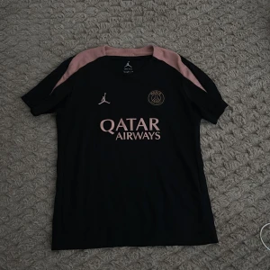 Svart PSG fotbollströja Jordan - Snygg svart PSG fotbollströja från Jordan med rosa detaljer på axlarna och rosa logotyper. Tröjan har korta ärmar och är tillverkad i ett lätt och ventilerande material, perfekt för träning eller match. Qatar Airways tryck på bröstet.