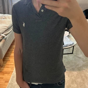 Mörkgrå pikétröja Polo Ralph Lauren S - Säljer en mörkgrå pikétröja från Polo Ralph Lauren i slim fit. Tröjan har korta ärmar, klassisk krage och två knappar framtill. Ikoniska vita Polo-loggan broderad på bröstet. Skön bomullskvalitet och stilren look. Priset kan såklart diskuteras 