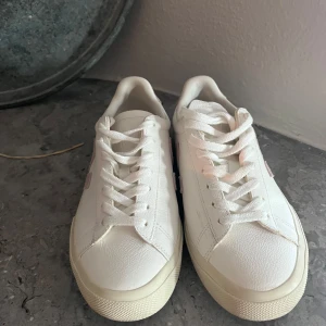 Veja vita sneakers med rosa detaljer - Säljer ett par fräscha Veja sneakers i vitt skinn med ljusrosa mockadetaljer på sidan och hälen. Klassisk låg modell med rund tå, platt sula och snörning. Perfekt för dig som gillar stilrena och trendiga sneakers med en clean vibe.