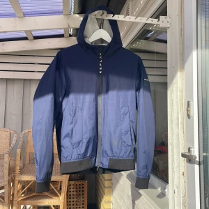 Blå vindjacka med huva från Replay - Snygg marinblå vindjacka från Replay med huva och svart ribbad kant vid ärmslut och nederkant. Jackan har dragkedja framtill, två stora fickor och vit meshfoder på insidan. Perfekt för dig som gillar stilren och sportig look.