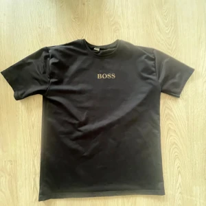 Svart BOSS t-shirt med tryck - Svart t-shirt från BOSS med guldigt logotryck på bröstet och en stor grafisk print med BOSS-text på ryggen. Klassisk rund hals och korta ärmar. Tillverkad i mjuk bomull för en skön känsla och snygg passform. Riktigt stilren!