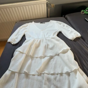 Vit volangklänning från 157 - Superfin vit långklänning från 157 med v-ringning, trekvartsärmar och flera lager volanger. Klänningen har en lätt rynkad struktur i tyget och en flowy passform som känns både luftig och trendig. Perfekt för sommarens alla tillfällen!