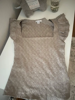Beige topp med volangärmar från AMISU - Supersöt beige topp från AMISU med fyrkantig ringning och volangärmar. Toppen har en vågig struktur i tyget och är tillverkad i ett mjukt material som känns skönt mot huden. Perfekt för dig som gillar en feminin och trendig stil.