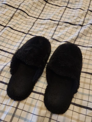 Svarta fluffiga tofflor - Mysiga svarta tofflor med fluffig yta och öppen häl. De har en mjuk ovandel och sula i gummi för extra komfort. Perfekta att glida runt i hemma när du vill ha det extra bekvämt. Storlek 39/40. Köpta på Ica för länge sen, de är orginelt en plast guld kedja som jag klippt av men det syns inte alls och dem är tvättade regelbundet. Inga som helst defekter.