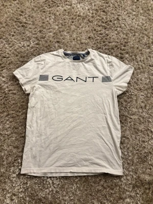 Vit GANT t-shirt med logga - Snygg vit t-shirt från GANT med klassisk logga i svart framtill och stilrena ränder på sidorna av trycket i storlek S.T-shirten har rund halsringning och är tillverkad i mjuk bomull. Perfekt för en clean och enkel stil. Har du några funderingar så skriv gärna🙏