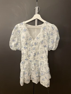 Vit blommig kortklänning med puffärm - Söt vit kortklänning med blått blommönster och v-ringning. Klänningen har puffärmar, volangdetaljer och en feminin siluett. Perfekt för varma dagar och somriga tillfällen.