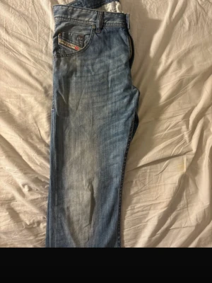 Blå raka jeans från Diesel - Säljer ett par klassiska blå jeans från Diesel med raka ben och snygg slitning framtill. Jeansen har fem fickor, knappgylf och detaljerade sömmar. Perfekta för en avslappnad och trendig look.