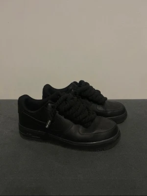 Nike Air force - Säljer ett par helsvarta Nike sneakers med chunky, rep-liknande svarta skosnören. Skorna har en klassisk låg siluett och är tillverkade i slätt läder med perforerade detaljer på tån. Perfekta för dig som gillar en clean och stilren look.