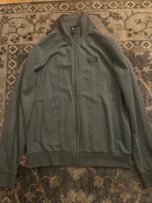 Grå ziptröja från EA7 Emporio Armani - Snygg grå ziptröja från EA7 Emporio Armani med hög krage och diskret logga på bröstet. Tröjan har två fickor framtill  och är tillverkad i ett mjukt material som känns skönt mot huden.