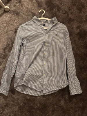 Randig skjorta från Ralph Lauren - Klassisk blåvit randig skjorta från Ralph Lauren med broderad logga på bröstet. Skjortan har lång ärm, knappar framtill och krage. Perfekt för en stilren look och tillverkad i mjuk bomull.