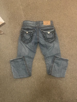 Blå True Religion jeans med kontrastsömmar - Snygga blå jeans från True Religion med tydliga kontrastsömmar och stora fickor bak med lock och knappar. Jeansen har en lätt sliten look framtill och klassisk femficksdesign. Perfekta för dig som gillar streetstyle och detaljer.