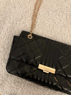 Svart axelväska Anna Field - Snygg svart axelväska från Anna Field med mönster och guldfärgade kedjor som axelrem. Väskan har en rektangulär form och ett elegant metallspänne framtill med logotyp. Materialet är glansigt konstläder. Bredd 28cm, djup 5cm, höjd 17cm.