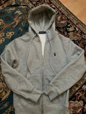 Grå zip-hoodie  - Snygg grå hoodie med dragkedja från Polo Ralph Lauren. Klassisk design med huva och dragsnören, samt den ikoniska blå broderade loggan på bröstet. Mjuk bomullsblandning och ribbade muddar för extra komfort. Storlek S men en liten S närmare xs hållet.