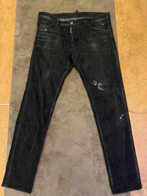 Svarta DSQ2 jeans med slitningar - Säljer ett par svarta jeans från DSQ2 med coola slitningar och distressed detaljer framtill. På baksidan finns DSQ2-loggan i orange på ena bakfickan. Jeansen har klassisk femficksdesign och raka ben. Perfekta för en trendig streetstil.        Kvitto finns från johnells inköpta 2023 men knappt använda då dom var förstora 
