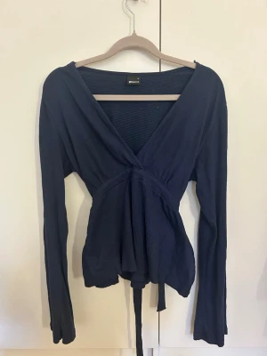 Mörkblå topp från Gina Tricot - Snygg mörkblå omlottopp från Gina Tricot med långa ärmar och knytband i midjan. Toppen har en djup v-ringning och är tillverkad i ett mjukt, lätt ribbat material som ger en skön passform. Perfekt att styla med jeans eller kjol.