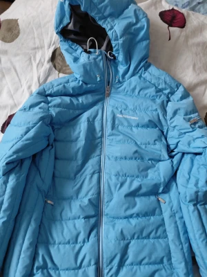 Blå dunjacka från Peak Performance - Snygg blå dunjacka från Peak Performance med huva och dragkedja framtill. Jackan har två sidofickor med dragkedja och diskret logga på bröstet. Perfekt för kalla dagar tack vare det värmande dunmaterialet och den sportiga designen.