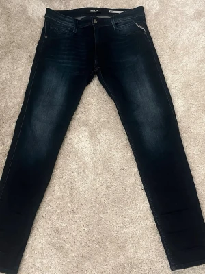 Mörkblå slim fit jeans från Replay - Snygga mörkblå slim fit jeans från Replay med diskreta slitningar och klassisk femficksdesign. Jeansen har en lätt tvättad look och smal passform som ger en modern siluett. Tillverkade i stretchigt denim för extra komfort. 