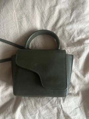 Grön handväska från ATP Atelier - Stilren grön handväska från ATP Atelier i den mindre storleken! Väskan har ett unikt lock med magnetlås och ett runt handtag samt axelrem. Perfekt för dig som gillar minimalistisk design och snygga detaljer. Väskan är rätt använd därav pris, dustbag tillkommer!❤️
