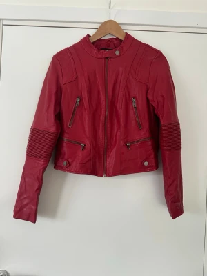 Röd skinnjacka med dragkedjor - Snygg röd skinnjacka med coola detaljer som dragkedjor framtill och vid ärmsluten. Jackan har ribbade partier på ärmarna och en hög krage med tryckknapp. Perfekt för dig som vill sticka ut med en färgstark look. Mycket gott skick!