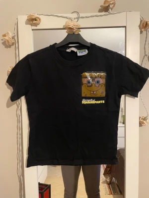 Svart Spongebob T-shirt - Cool svart t-shirt med Spongebob Squarepants-tryck på bröstet och stor vit Spongebob-grafik på ryggen. Klassisk passform och rund halsringning. Perfekt för dig som älskar tecknade serier och vill sticka ut med en unik t-shirt.