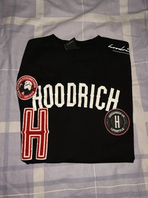 Svart Hoodrich t-shirt med patches - Snygg svart t-shirt från Hoodrich med stora vita bokstäver på bröstet och flera broderade patches i rött, vitt och svart. T-shirten har en stor röd H, rund patch med texten 'Nothing to something' och en rund Hoodrich UK Lifestyle patch. Skön passform och streetwear-vibe.
