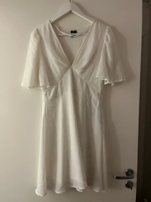 Vit klänning med v-ringning - Säljer en vit klänning med v-ringad hals och fladdriga korta ärmar. Klänningen har en lätt utsvängd passform och är tillverkad i ett tunt, mjukt material som ger ett luftigt intryck. Andvänt en gång storlek M men passar som S