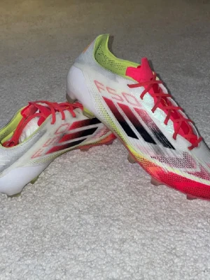 Adidas vita och röda fotbollsskor - Säljer ett par snygga Adidas fotbollsskor med vit bas, röda och gula detaljer samt svarta ränder på sidan. Skorna har röda snören och en låg profil för bra bollkontroll. Ovandelen är i syntetmaterial och sulan är anpassad för spel på gräs.