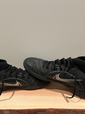 Svarta Nike indoor football shoes  - Säljer ett par svarta Nike fotbollsskor med vit swoosh och texten 'NIKE FOOTBALL' på sidan. Skorna har snörning, låg profil och strumpliknande skaft för extra stöd. Yttermaterialet är syntetiskt och designen är sportig och modern.