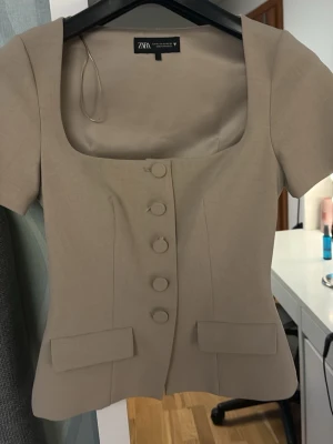Beige topp med knappar från Zara - Snygg beige topp från Zara med korta ärmar och markerad fyrkantig ringning. Toppen har dekorativa klädda knappar framtill och två fickdetaljer. Figurnära passform och stilren design, perfekt för att lyfta vilken outfit som helst. I super fin skick ända är att det är lite trådar vid knapparna som man ser nära på tredje bilden🥰🥰ändast använd en gång 