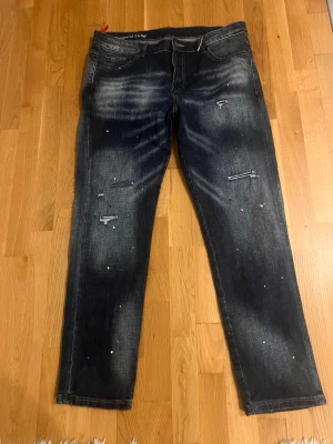 Mörkblå distressed jeans med stänk - Säljer ett par mörkblå jeans med slitningar och vita färgstänk för en cool, avslappnad look. Jeansen har raka ben, klassisk femficksmodell och råa benslut. Materialet är denim i bomull. Perfekta för dig som gillar streetwear och en trendig stil.