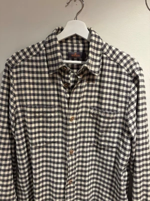 Rutig skjorta från Morris - Snygg rutig skjorta från Morris i svart och vit med klassisk krage och bröstfickor. Skjortan har långa ärmar och knäppning framtill med bruna knappar. Perfekt för en avslappnad men stilren look.