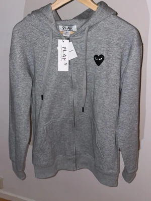 Grå hoodie från Comme des Garçons Play - Grå zip-hoodie med svart hjärta och ögon-tryck på bröstet och stort svart hjärta med ögon på ryggen. Mjuk bomull, dragsko i huvan och fickor framtill. Perfekt för en chill och stilren look.