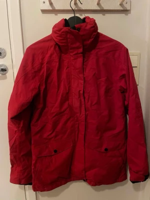 Fjällräven jacka - Snygg röd vindjacka med hög krage och två stora fickor framtill med knappar. Jackan har justerbara muddar med kardborre och stängs med dragkedja och tryckknappar. Perfekt för blåsiga dagar och enkel att matcha.