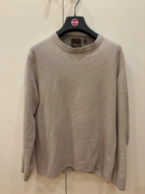 Beige stickad tröja från Oscar Jacobson - En stilren beige stickad tröja från Oscar Jacobson med rund halsringning och ribbade muddar vid ärmslut och nederkant. Perfekt för lager-på-lager och enkel att matcha med olika outfits.