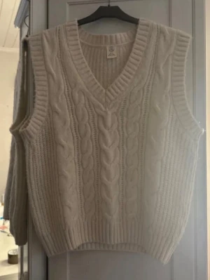 Vit kabelstickad väst med v-ringning - Kabelstickad väst i offwhite med v-ringning och breda ribbade kanter. Klassisk design med grovstickat mönster framtill och slätstickad baksida. Perfekt att styla över skjorta eller t-shirt.