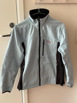 PeakPerformance Fleecejacka - Sportig ljusblå vindjacka från Peak Performance med svarta paneler på sidorna. Jackan har dragkedjor framtill och på fickorna samt hög krage. Perfekt för blåsiga dagar och utomhusaktiviteter.