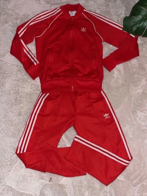 Rött Adidas loungewear-set - Säljer ett rött Adidas loungewear-set med klassiska vita ränder längs ärmar och ben. Setet består av zip-jacka och byxor, båda med Adidas logga framtill. Tillverkat i Primeblue-material med Parley Ocean Plastic. Perfekt för chill dagar hemma.