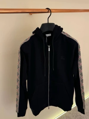 Svart Burberry hoodie med dragkedja - Snygg svart hoodie från Burberry med dragkedja framtill och huva med snören. Ärmarna har det klassiska Burberry-mönstret längs sidorna. Perfekt för en trendig och avslappnad stil. Mjukt material och två fickor framtill.