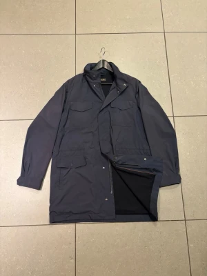 Mörkblå Traveller Coat från Loro Piana - Stilren mörkblå kappa från Loro Piana i modellen Traveller Coat. Kappan har fyra framfickor med lock, hög krage och dold dragkedja med knappslå. Insidan har en innerficka med orange kant. Perfekt för dig som vill ha en klassisk och funktionell look.