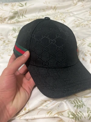 Svart Gucci keps med GG-mönster - Säljer en svart keps från Gucci med klassiskt GG-monogram över hela kepsen. På sidorna finns de ikoniska gröna och röda ränderna. Kepsen har böjd skärm och är tillverkad i ett slitstarkt tygmaterial. Perfekt för dig som vill ha en stilren och lyxig accessoar.