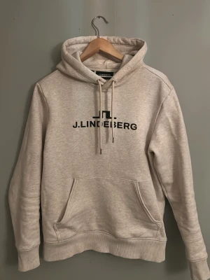 Beige hoodie från J.Lindeberg - Snygg beige hoodie från J.Lindeberg med svart logga på bröstet. Klassisk modell med huva, dragsnören och stor magficka. Tillverkad i mjukt material som känns skönt mot huden. Perfekt för en avslappnad och trendig look.