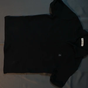 Svart polotopp från The Blue Uniform - Klassisk svart polotopp från The Blue Uniform i ribbad bomull. Tidlös design med korta ärmar och liten krage. Perfekt att matcha med jeans eller kjol för en clean look.