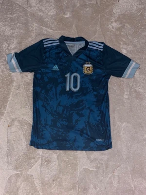 Argentina Messi matchtröja blå - Säljer en blå Argentina fotbollströja från Adidas med nummer 10 och Messi på ryggen. Tröjan har ljusblå detaljer, korta ärmar och AFA-emblem på bröstet. Tillverkad i lätt och ventilerande material med Aeroready-teknologi.