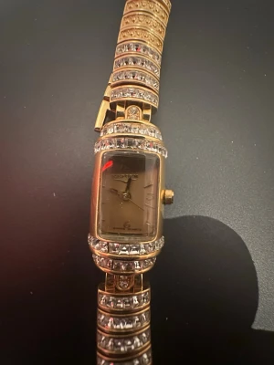 Guldig rektangulär klocka med stenar - Snygg guldig klocka från Geneva med rektangulärt ur och armband dekorerat med glittriga stenar. Klockan har en lyxig känsla och detaljerade mönster på armbandet. Perfekt accessoar för att sticka ut och ge outfiten extra bling. Funkar ej!