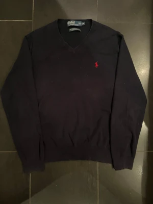 Mörkblå v-ringad tröja Polo Ralph Lauren - Snygg mörkblå v-ringad tröja från Polo Ralph Lauren i 100% pima bomull. Klassisk design med röd broderad logga på bröstet och ribbade muddar vid ärmslut och nederkant. Perfekt för en stilren och tidlös look.