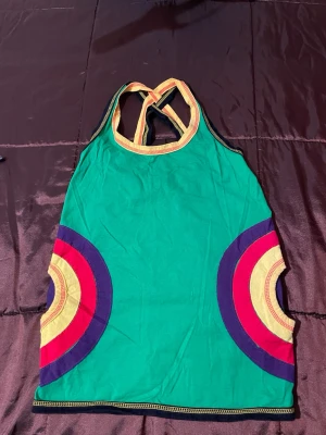 Grön linnetopp med färgglada detaljer - Snygg grön linnetopp med brottarrygg och färgglada cirkeldetaljer i lila, rött och gult på sidorna. Axelbanden är randiga i gult och grönt för en sportig vibe. Perfekt för dig som gillar färg och unik stil..storlek smal