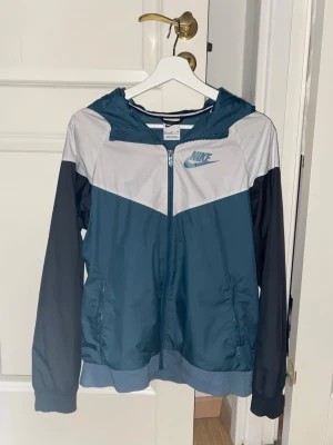 Nike jacka blå vit svart - Nike vindjacka i blått, vitt och svart med dragkedja framtill och huva. Jackan har svarta ärmar, två fickor och en klassisk Nike-logga på bröstet. Tillverkad i lätt och tåligt material, perfekt för blåsiga dagar. Snygg färgblockdesign som ger en sportig look.