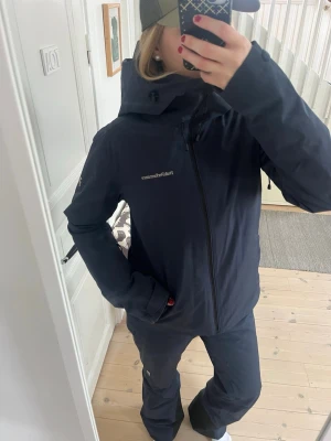Mörkblå skidkläder Peak Performance - Säljer detta snygga set med skidkläder i modellen Anima från peak performance i marinblå. De är medium vadderade och tillverkade i ett vattentätt HIPE material. Byxorna går ner till marken på mig som är 168cm och kan spännas åt i midjan. Byxorna har lite slitningar längst ner på innerbenet (se bild). Jackan har en stor luva som går över hjälmen, ventilation under armarna och fickor med dragkedja  Storleken är M men passar mig som brukar ha S och är 168 cm lång.  Ny pris 6500.