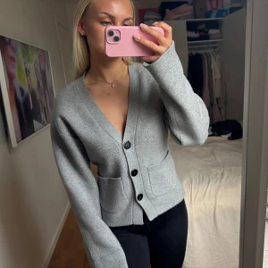 Grå kofta med knappar och fickor - Snygg grå kofta med v-ringning och tre svarta knappar framtill. Koftan har två praktiska fickor och är i ett mjukt, stickat material. Perfekt för lager-på-lager och chill vardagsstil.